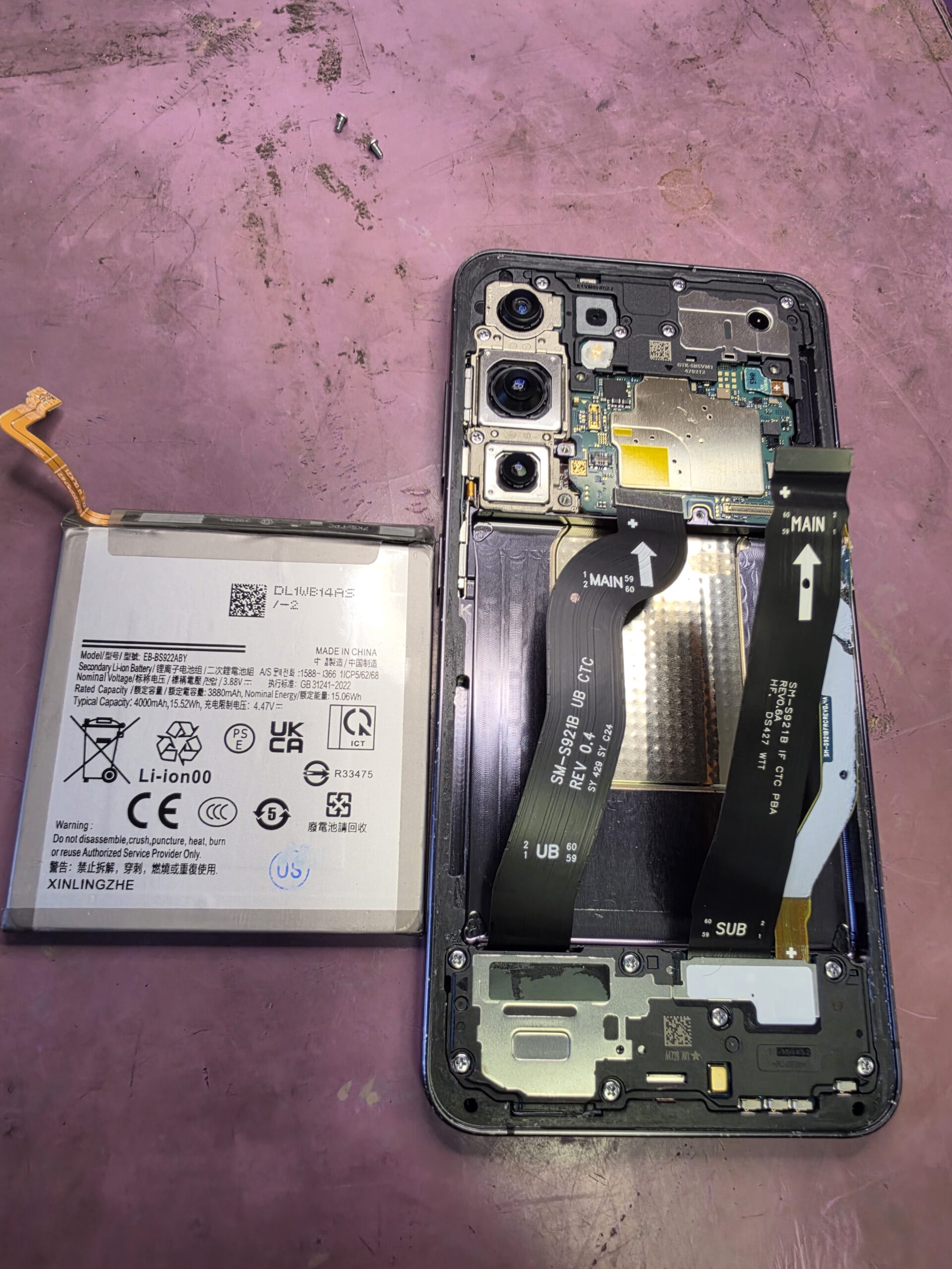 Removed non-genuine replacement battery beside the opened Samsung Galaxy S24, with its empty battery compartment and ribbon cables visible