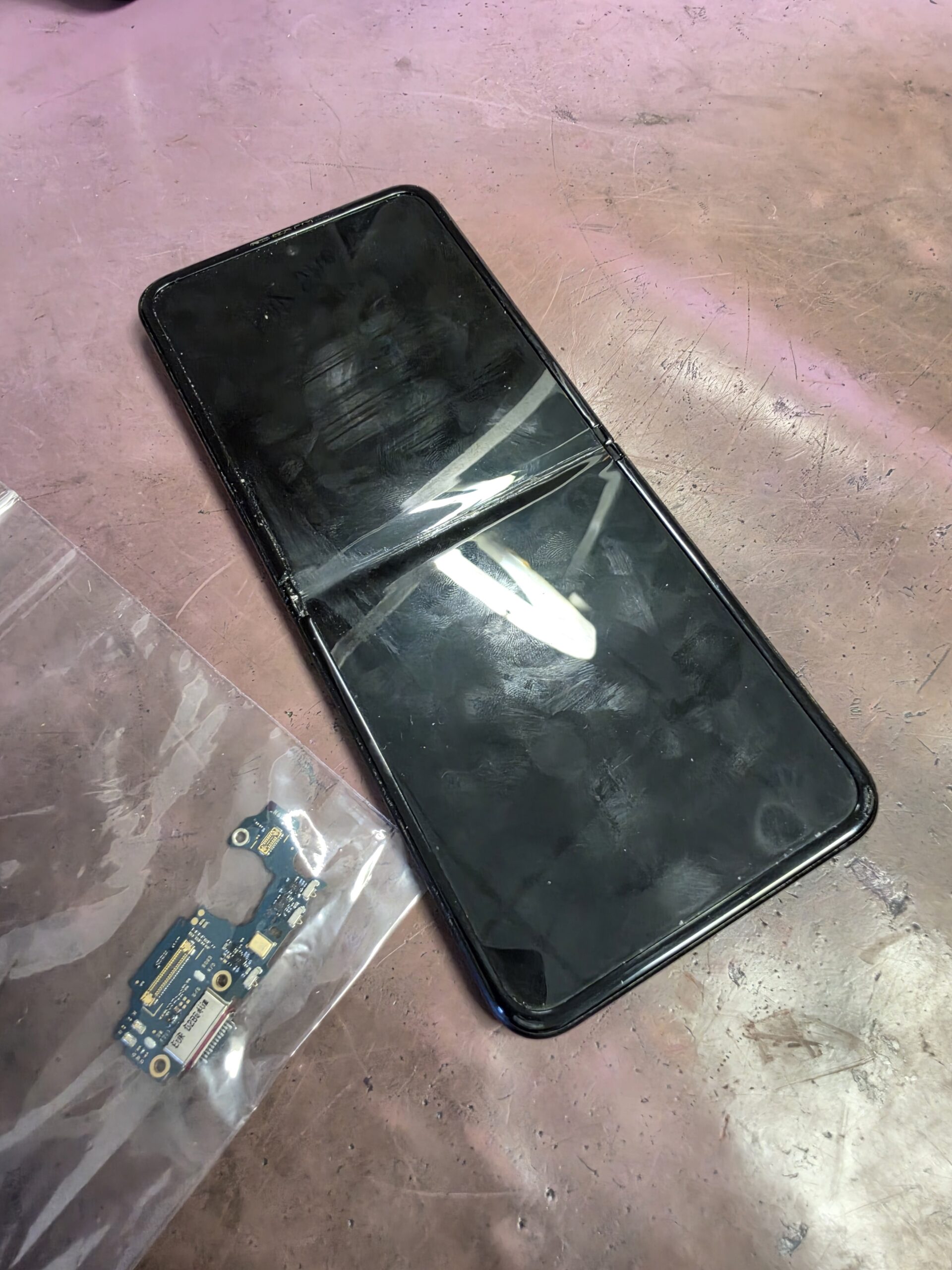 Samsung Galaxy Flip on the workshop bench beside a bagged charging-port circuit board