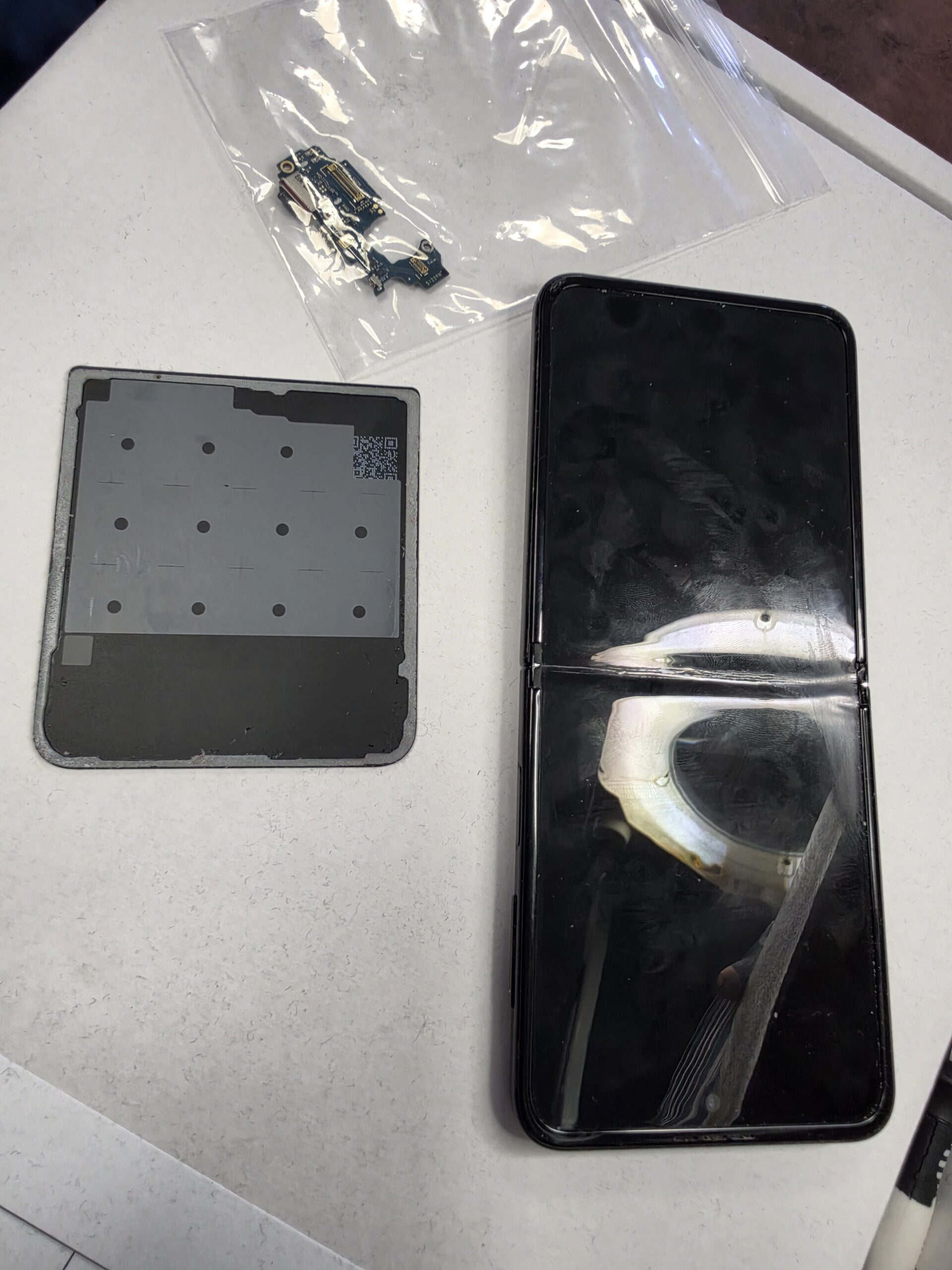 Samsung Galaxy Flip face-up beside its removed rear panel and a bagged circuit board during workshop repair